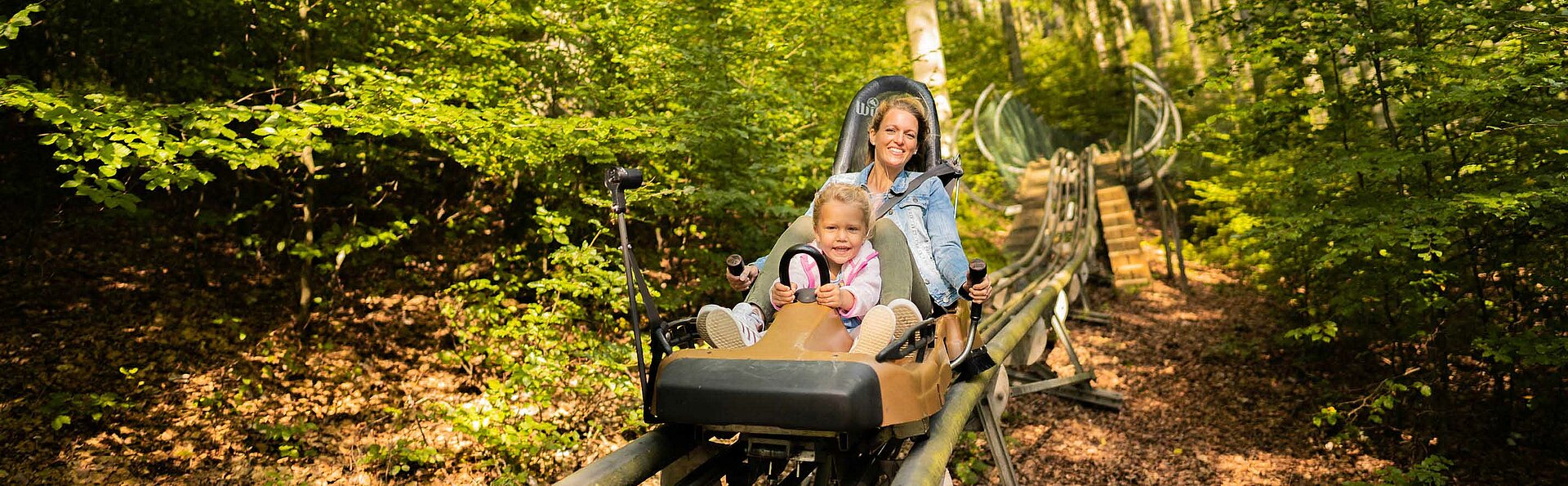 Die Mutter rast mit ihrer begeisterten kleinen Tochter mit der Sommerrodelbahn den Hang hinunter.